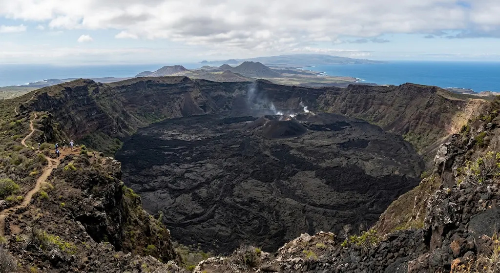 Panoramic view of the vast crater of the Sierra Negra Volcano on Isabela Island, Galápagos, with tourists on the rim and the Pacific Ocean in the background.